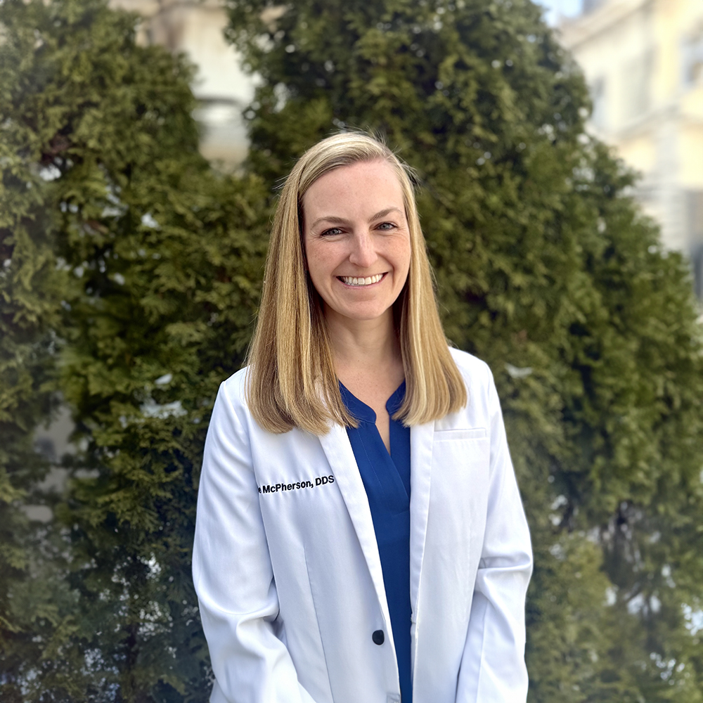 The image shows a woman in a white lab coat standing outdoors against a backdrop of greenery and a building, smiling at the camera.