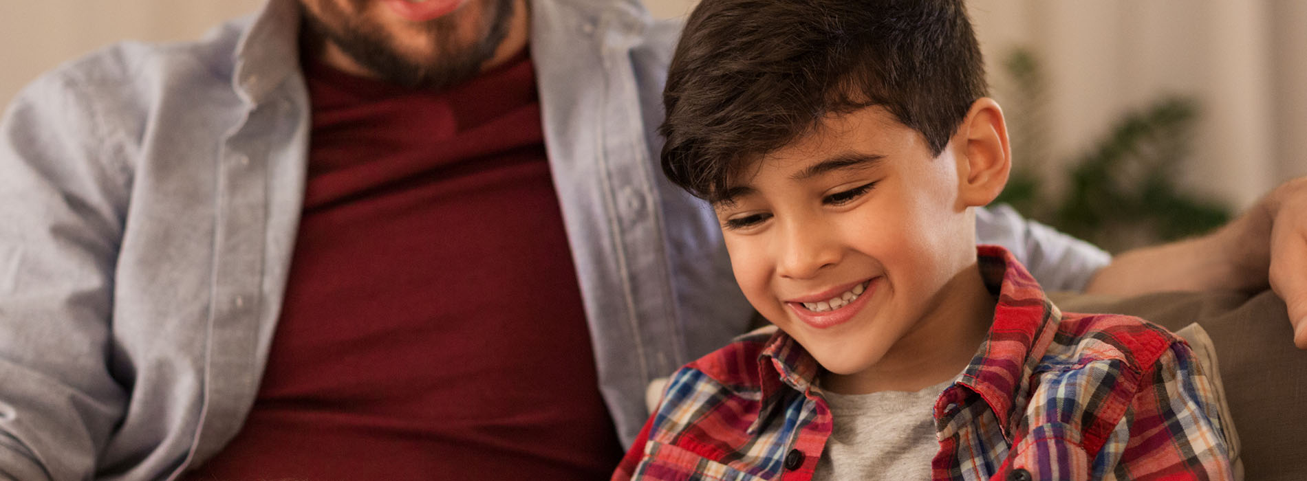 The image shows a man and a child sitting together on a couch, both smiling at the camera, with the man wearing a plaid shirt.