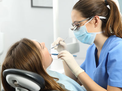 The image shows a dental professional performing a procedure on a patient while seated at a dental chair, with both individuals wearing personal protective equipment.